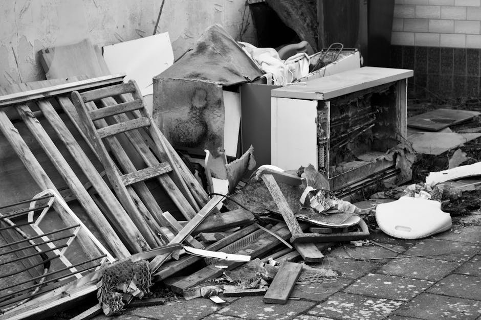 A pile of discarded household furniture and waste materials outside a property, including a wooden garden bench with slats, a broken cupboard or television stand with a damaged door, a foam toilet seat, and various cardboard boxes and packaging debris. Items are stacked against an exterior wall with some on the pavement; a portion of a brick wall and concrete ground are visible in the background. The scene appears cluttered with construction or disposal waste, with materials such as wood, plastic, and fabric present. This image illustrates the type of bulky waste that may require professional removal services, like those offered by Man with Van Willesden, during home relocation or clearance projects, aligning with the context of adhering to Brent Council's waste disposal rules.