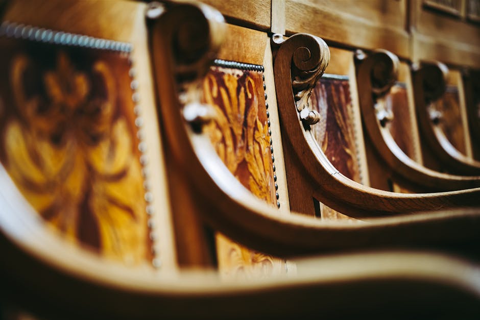 Close-up image of ornate wooden chairs with carved armrests and decorative paneling on the backs, featuring intricate woodgrain patterns and polished surfaces. The chairs are arranged in a row indoors, with visible spacing between each piece. The image captures the detailed craftsmanship of the furniture, which appears to be part of a large wooden dining set or similar collection. The lighting highlights the natural finish of the wood, emphasizing the smooth curves and fine detailing. This setting relates to home furniture or packing during a household move, as part of furniture transport and home relocation processes. Man with Van Willesden, a professional removals service, specializes in packing and transporting such items during house removals, ensuring careful handling of valuable furniture for clients in the WILLESDEN area. The focus on furniture details supports the theme of moving logistics and furniture transport within the context of managing household contents in compliance with local waste rules, such as Brent Council bulky waste regulations.