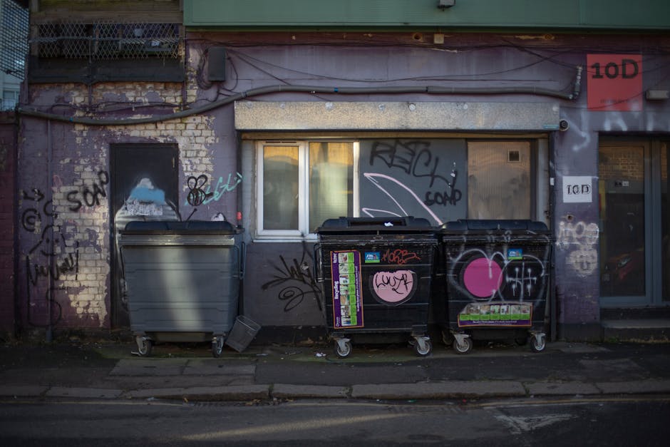 A pile of discarded household furniture and waste materials outside a property, including a wooden garden bench with slats, a broken cupboard or television stand with a damaged door, a foam toilet seat, and various cardboard boxes and packaging debris. Items are stacked against an exterior wall with some on the pavement; a portion of a brick wall and concrete ground are visible in the background. The scene appears cluttered with construction or disposal waste, with materials such as wood, plastic, and fabric present. This image illustrates the type of bulky waste that may require professional removal services, like those offered by Man with Van Willesden, during home relocation or clearance projects, aligning with the context of adhering to Brent Council's waste disposal rules.
