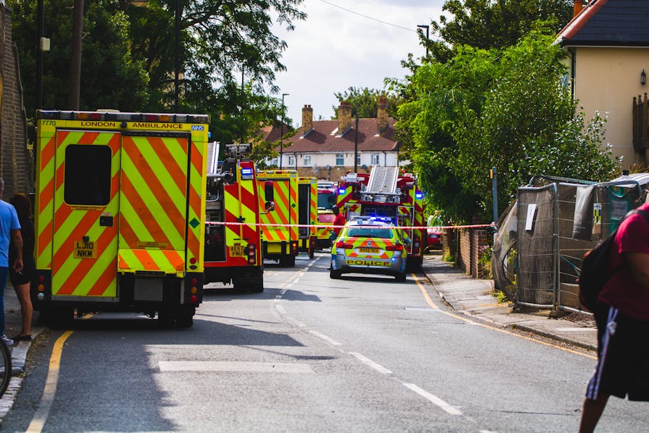 A scene of an emergency response on a residential street, featuring multiple fire trucks with yellow and red chevron markings parked along the road, and a police car with flashing blue lights positioned nearby to manage the situation. The fire trucks are equipped with ladders, hoses, and other firefighting tools, and are arranged in a line, blocking part of the street to ensure access for emergency services. To the right, there is a construction or storage area enclosed by a makeshift fence made of black plastic sheeting and wooden posts. The background shows houses with chimneys, trees, and overhead power lines under daytime lighting conditions. Pedestrians are seen on either side of the road, observing the scene, which appears to involve emergency responders potentially involved in a home-related incident or safety operation, supported by [COMPANY_NAME] for logistics or scene management in the context of house removals or relocations.
