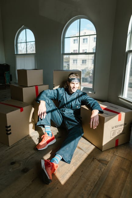 A young man with long hair, wearing a blue work jumpsuit, red sneakers, and striped socks, is sitting on a wooden floor inside a well-lit room with large arched windows. He is leaning against a stack of cardboard moving boxes secured with red packing tape, some of which are placed directly on the floor while others are stacked behind him. The sunlight streaming through the windows creates shadows on the floor, highlighting the packing materials and the man's relaxed posture. The scene captures the stages of a home relocation process managed by Man with Van Willesden, with visible objects indicating a packing and moving environment, such as boxes ready for transport. The room appears clean and minimally furnished, suggesting an active moving or packing phase as part of a furniture transport or house removals service.