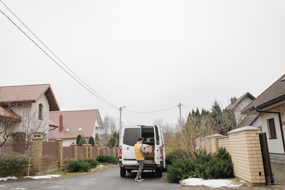A man wearing a yellow jacket and black hat is seen loading a cardboard box into the back of a white van parked on a residential street in Willesden, NW10. The van is positioned near a house with a br