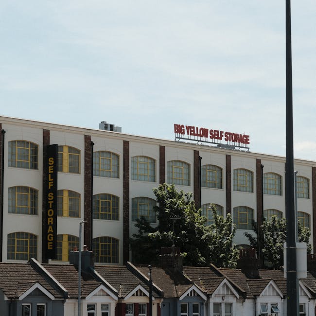 Exterior view of a three-storey commercial building with a white facade and brown brick accents, featuring large, evenly spaced yellow-framed windows on each level. On the left side of the building, a vertical black sign reads 'SELF STORAGE' in yellow letters. Atop the building, a red-lit sign displays 'BIG YELLOW SELF STORAGE'. In front of the building, there are small residential-style houses with tiled roofs and white window frames, partially obscured by green trees and shrubs. A tall street lamp and a utility pole are visible in the foreground, with the sky above appearing partly cloudy. The scene suggests a business involved in home relocation and furniture transport, relevant to the context of removals and packing services offered by Man with Van Willesden.