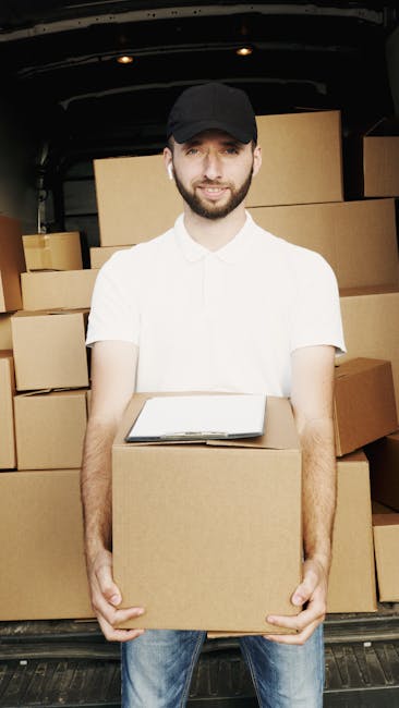 A man wearing a black cap, white polo shirt, and jeans stands inside the back of a van, holding a medium-sized cardboard box at waist height. The van is filled with multiple stacked cardboard boxes, some sealed with packing tape, arranged in an organized manner. The background shows the interior of the van, with a dark ceiling and sides. The scene depicts a home relocation or furniture transport process, with the man assisting in packing, loading, or moving boxes as part of a professional removals service provided by Man with Van Willesden, supporting efficient packing and moving logistics.