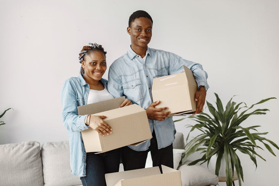 An indoor scene showing two individuals during a house relocation process in a kitchen. A woman with long dark hair wearing a white t-shirt is smiling and holding a small cardboard box, while a man wi