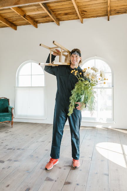 A man with light skin, wearing a black long-sleeved shirt, dark pants, and bright orange sneakers, is standing inside a well-lit room with wooden flooring and a wooden ceiling with exposed beams. He is smiling and holding a stack of two wooden chairs with one hand, balanced on his shoulder. In his other hand, he carries a large bouquet of mixed flowers and greenery. The room features two arched windows allowing natural light to fill the space, with a green upholstered armchair visible on the left side. The scene depicts the home relocation process, involving furniture transport and packing, with the man representing a professional removals service by Man with Van Willesden, supporting the theme of house removals and moving logistics.