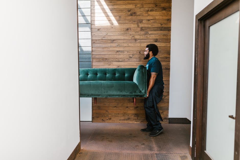 A man with a beard and wearing a dark blue uniform is seen inside a residential property, carefully lifting a green upholstered sofa with wooden legs through a doorway. The sofa is partially wrapped in protective green fabric, indicating it is being prepared for a home relocation or furniture transport. The background features a wooden feature wall with horizontal planks, and the floor is covered with a brown carpet. The doorway opens to a corridor with white walls and a wooden door frame on the right. This scene demonstrates the process of packing, loading, or moving furniture as part of a professional removals service offered by Man with Van Willesden, located near Roundwood Park in Willesden, highlighting typical furniture handling and loading procedures involved in house removals.