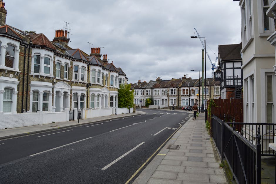 A quiet residential street in Willesden with a row of Victorian-style terraced houses featuring white stucco facades, bay windows, and red tiled roofs under an overcast sky. The pavement runs alongside the houses, bordered by a black metal fence and some greenery. A large blue and white vehicle, likely a van used by Man with Van Willesden, is parked on the street, with its rear doors open for loading or unloading furniture and packing materials. Nearby, several cardboard boxes, wrapped furniture, and packing blankets are visible on the pavement, indicating a home relocation or moving process. A person is seen walking along the sidewalk, and the scene includes street lamps and a construction crane in the distance, suggesting ongoing urban development. The overall scene captures a typical UK house move setup, with details supporting furniture transport and moving logistics for a NW10 flat move, aligned with house removals services provided by Man with Van Willesden.
