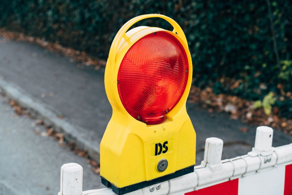 A large yellow warning light with a prominent red circular reflector on its front, mounted on top of a portable traffic barrier. The barrier is positioned on a paved road next to a curb, with white fe