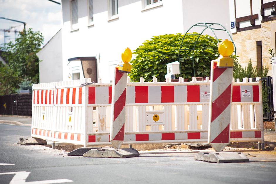 A large yellow warning light with a prominent red circular reflector on its front, mounted on top of a portable traffic barrier. The barrier is positioned on a paved road next to a curb, with white fe