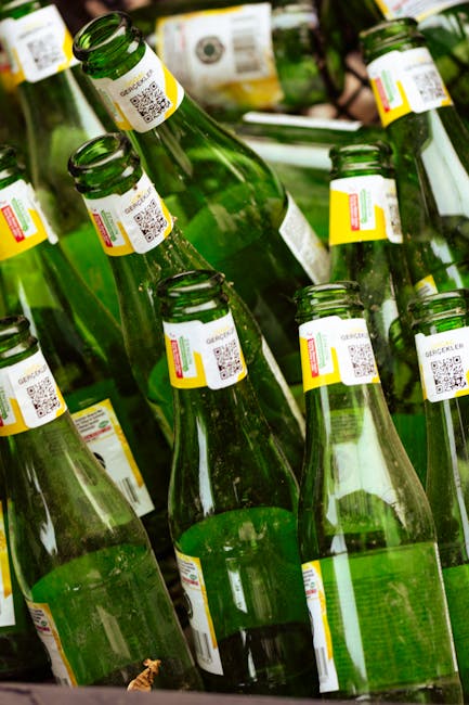 A collection of green glass bottles with white and yellow temperature-sensitive labels featuring QR codes, stacked and arranged closely together outdoors. The bottles are placed on a surface, and some are leaning against each other. The background includes blurred natural elements such as plants and foliage, suggesting a recycling or disposal process possibly tied to eco-friendly packing materials used during house removals. This scene reflects environmentally conscious practices related to furniture and household item disposal, as part of a home relocation or moving service provided by Man with Van Willesden.