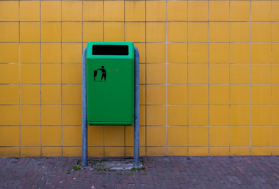 A green public waste bin with a black pictogram of a person disposing of rubbish, mounted on a metal stand against a yellow tiled wall outside a building. The bin has a black rectangular opening at the top for waste disposal and a small red light or indicator. The ground is paved with red bricks, and there are some small patches of dirt and grass at the base of the stand. This scene may be part of a building's external area where waste is managed, relevant to home relocation or moving logistics services offered by Man with Van Willesden, such as arranging disposal or recycling of items during furniture transport or packing and moving activities.