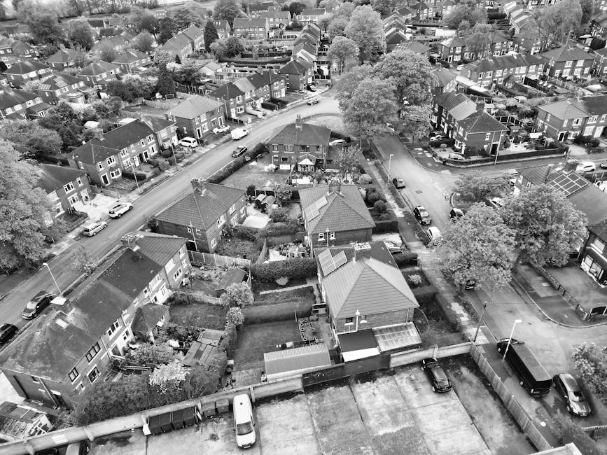 An aerial black-and-white photograph shows a residential neighbourhood with rows of terraced houses and detached homes, some with solar panels on their roofs. The image captures a street with parked cars along both sides, winding through the community. Several gardens with trees, hedges, and lawns are visible, along with fences delineating private yards. Near the bottom of the image, a driveway leads to a parking area with a few vehicles, adjacent to a property with a small garden and a shed. The surrounding environment includes a mix of paved roads, sidewalks, and green spaces, with mature trees providing shade and greenery throughout the area. The scene suggests a typical suburban setting, with potential for home relocation or furniture transport activities coordinated by local moving companies such as Man with Van Willesden, highlighting the importance of permits managed by Brent Council for effective logistics during house moves.