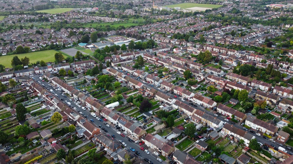 An aerial view of a residential neighbourhood showing rows of terraced houses with red and brown tiled roofs, arranged along a network of streets and pavements. Many houses have small front gardens, some with visible grass lawns, plants, and trees. Cars are parked along the roads and in driveways, with some in the process of being loaded onto or unloaded from a large white moving van parked on the street. The van is positioned adjacent to the pavement near an open doorway of a house, with a person visible carrying a cardboard box into the vehicle, supported by a trolley to facilitate the home relocation process. The image captures the typical layout of suburban houses, with a few larger green spaces and playing fields visible in the background, illuminated by daylight and natural outdoor lighting. This scene exemplifies the logistics involved in house removals and furniture transport within the Willesden area, supported by professional services like those provided by Man with Van Willesden.
