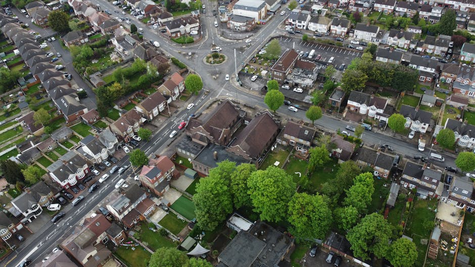 An aerial black-and-white photograph shows a residential neighbourhood with rows of terraced houses and detached homes, some with solar panels on their roofs. The image captures a street with parked cars along both sides, winding through the community. Several gardens with trees, hedges, and lawns are visible, along with fences delineating private yards. Near the bottom of the image, a driveway leads to a parking area with a few vehicles, adjacent to a property with a small garden and a shed. The surrounding environment includes a mix of paved roads, sidewalks, and green spaces, with mature trees providing shade and greenery throughout the area. The scene suggests a typical suburban setting, with potential for home relocation or furniture transport activities coordinated by local moving companies such as Man with Van Willesden, highlighting the importance of permits managed by Brent Council for effective logistics during house moves.