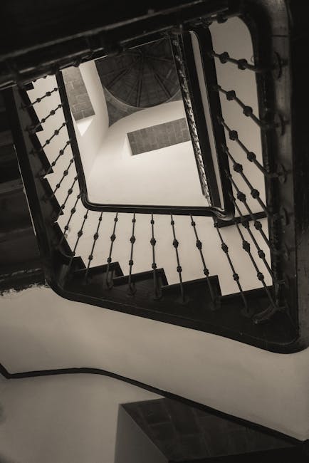A black metal spiral staircase viewed from the top looking downward, with vertical balustrades and evenly spaced balusters, inside a white-walled building. The staircase's steps are dark-colored, likely wood or metal, and the surrounding walls are painted white, providing a contrast. The image captures the open central void of the staircase, showing multiple landings as the staircase winds around a central axis. This interior environment may be part of a residential or commercial property undergoing home relocation or moving preparations, with the staircase possibly serving as a route for moving furniture and boxes between floors. Man with Van Willesden often handles such tasks involving navigating narrow staircases during furniture transport and packing and moving services, as suggested by the contextual link to house removals and moving logistics.