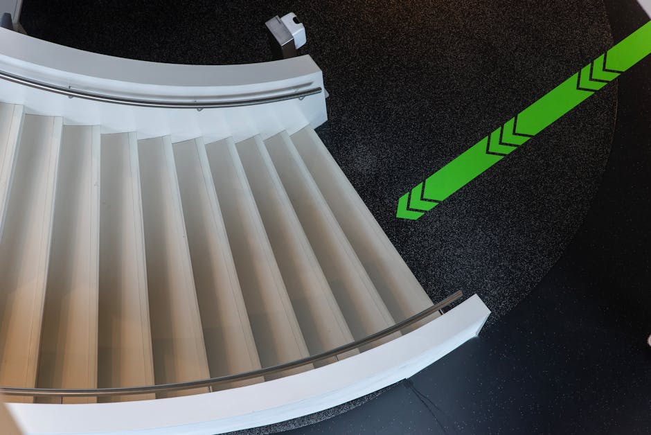 A top-down view of a curved staircase inside a property, featuring white steps with a smooth surface and a metallic handrail along the outer edge. The staircase spans multiple levels, with the steps smoothly transitioning from one floor to another. Adjacent to the staircase, the dark flooring is visible, textured with small, reflective specks that suggest a polished finish. To the right, bright green arrow markings are painted on the floor, indicating directional guidance for safe navigation or moving routes. The lighting is even, illuminating the entire scene clearly, which facilitates visual clarity for viewers with accessibility needs. The context appears to be part of an internal home environment, likely being prepared for a house removal or furniture transport, a service offered by Man with Van Willesden, as indicated by the webpage information. This setting highlights the importance of careful planning and the use of appropriate equipment for interior furniture transport or home relocation activities.