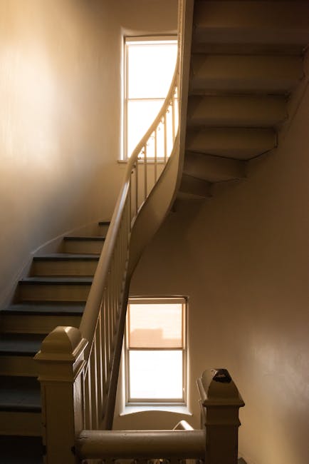 A black metal spiral staircase viewed from the top looking downward, with vertical balustrades and evenly spaced balusters, inside a white-walled building. The staircase's steps are dark-colored, likely wood or metal, and the surrounding walls are painted white, providing a contrast. The image captures the open central void of the staircase, showing multiple landings as the staircase winds around a central axis. This interior environment may be part of a residential or commercial property undergoing home relocation or moving preparations, with the staircase possibly serving as a route for moving furniture and boxes between floors. Man with Van Willesden often handles such tasks involving navigating narrow staircases during furniture transport and packing and moving services, as suggested by the contextual link to house removals and moving logistics.