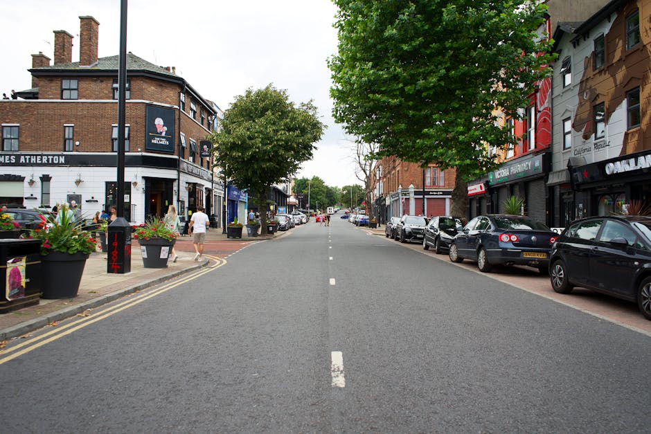 A straight urban street scene during daytime with a row of parked cars on the right side, including sedans in dark colours, and a few pedestrians walking along the sidewalk on the left side. The sidewalk features large decorative planters with blooming flowers, and a black lamppost standing near a double yellow line marking the edge of the street. On the left, a corner building with a rounded facade houses a bar or cafe, with outdoor seating and windows displaying signs. The buildings on both sides display a mix of brick and painted facades, with signage indicating various shops and businesses. Green trees with dense foliage line parts of the street, providing shade, while the roadway has a single dashed white line down the center. The scene is lit with natural daylight, capturing the typical appearance of a busy commercial area in Willesden, suitable for house removals and furniture transport logistics, as handled by Man with Van Willesden.